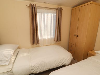 A small bedroom with two beds a window with curtains and a wooden wardrobe at Lodge 3 in Silverdale