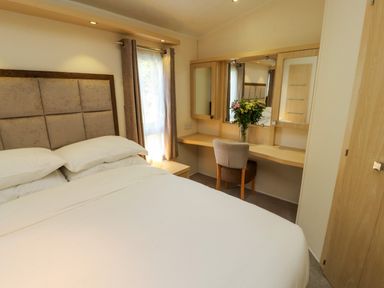 A bedroom with a bed pillows window curtains and a dressing table with chair and flowers at Lodge 3 in Silverdale