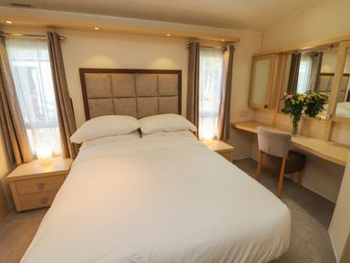 A bedroom with a double bed bedside tables windows curtains a desk chair and flowers at Lodge 3 in Silverdale