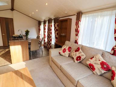 A living room with a beige sofa and floral cushions next to windows with matching curtains and a dining area with chairs near a kitchen counter at Lodge 4 in Silverdale