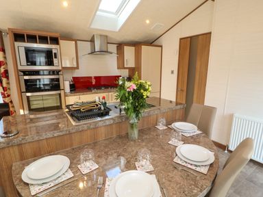 A dining area with a round table set for four with plates cutlery and glasses and a kitchen with appliances and a flower vase at Lodge 4 in Silverdale