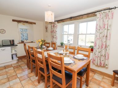 A dining room with a wooden table set for eight with plates and wine bottles by windows with floral curtains at High Wood Foot in Bampton Cumbria