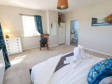 A bedroom with a bed featuring folded towel animals and pillows a wooden chair near a window with blue curtains a white chest of drawers and an en suite bathroom at High Wood Foot in Bampton Cumbria