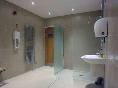 A bathroom with a wall-mounted hand dryer a sink a soap dispenser a towel radiator a wooden door and a frosted glass shower partition at 6 Burgh Island in Bigbury-On-Sea