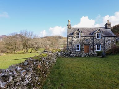A stone cottage with four windows and a wooden door surrounded by a stone wall in a grassy field at Tyn Y Graig in Fairbourne