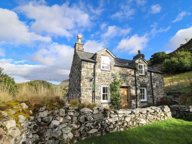 A stone house with a wooden door and windows surrounded by a stone wall and grass at Tyn Y Graig in Fairbourne