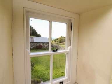 A window with white frame showing a green lawn stone building and trees outside at Tyn Y Graig in Fairbourne
