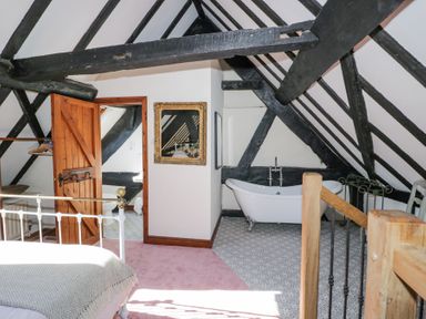 A bedroom with exposed black beams a white bed an open wooden door a bathtub and patterned floor at Wyre Court Cottage in Bewdley