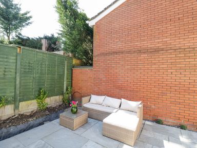 An outdoor patio with a wicker sofa and table with flowers against a brick wall and green fence at Wyre Court Cottage in Bewdley