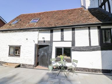 An outdoor patio with stone tiles a small round table with two chairs a vase with flowers and a bottle of wine outside a white and black timber framed building at Wyre Court Cottage in Bewdley