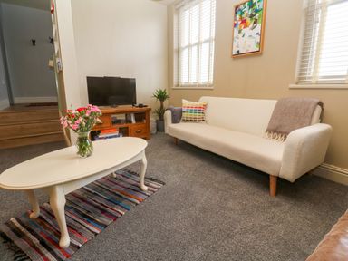 A living room with a white sofa a colorful cushion a coffee table with flowers and a TV on a wooden stand at Cottage Annex in Constantine