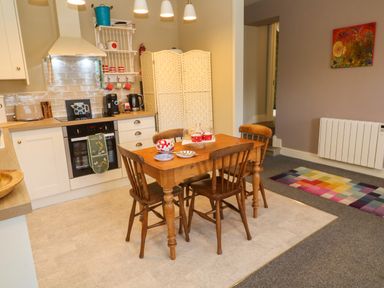 A kitchen with a wooden dining table and four chairs a white cabinet with a stove and oven a white shelf with cups a colorful rug and a painting on the wall at Cottage Annex in Constantine