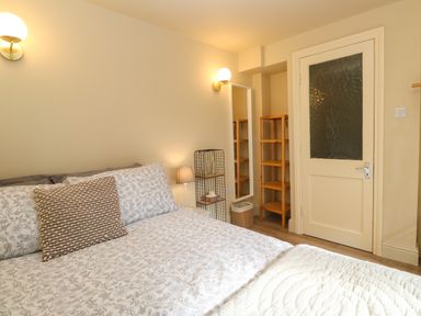 A bedroom with a bed with floral bedding a pillow wall lights a wooden shelf a full length mirror and a door at Cottage Annex in Constantine