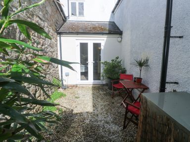 A small outdoor patio with gravel ground two red chairs a wooden table and plants near white double doors at Cottage Annex in Constantine