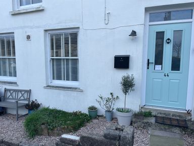 The exterior of a white house with a light blue door numbered 44 a black mailbox a bench and several potted plants in front of the Cottage Annex in Constantine