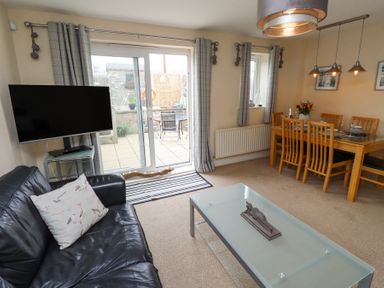 A living room with a black leather sofa and a glass coffee table next to a dining table with wooden chairs and a TV mounted near sliding doors at Sea Shore in Seahouses
