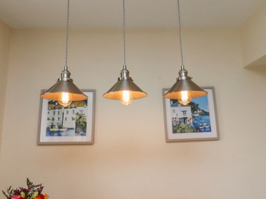 Three hanging metal light fixtures with exposed bulbs above two framed paintings on a beige wall at Sea Shore in Seahouses