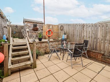 An outdoor patio area with a glass table and four chairs next to a wooden deck with steps and a small shed at Sea Shore in Seahouses