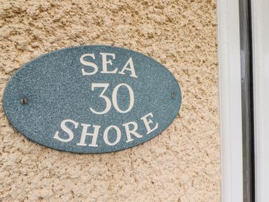 A blue oval house sign with white letters reading Sea 30 Shore on a textured beige wall at Sea Shore in Seahouses