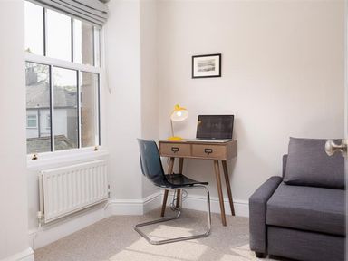A small room with a wooden desk laptop yellow lamp black chair gray sofa and a window at Bellflower Cottage in Windermere