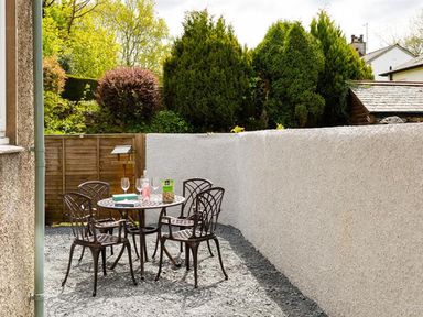 A small outdoor patio area with a round table and four metal chairs surrounded by a white wall and greenery at Bellflower Cottage in Windermere