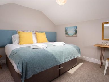 A bedroom with a large bed with yellow pillows and blue blanket a painting on the wall and a small wooden table with a mirror at Bellflower Cottage in Windermere