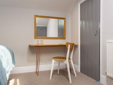 A small wooden desk with two bottles and a wooden chair in front of a wall mirror with a gold frame in a room with a grey door and beige carpet at Bellflower Cottage in Windermere