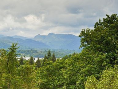 A landscape with trees in the foreground a lake and mountains in the distance under a cloudy sky at Bellflower Cottage in Windermere