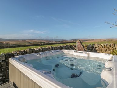 An outdoor hot tub next to a stone wall with fields and hills in the background at Petty Knowes Cottage in Rochester near Otterburn