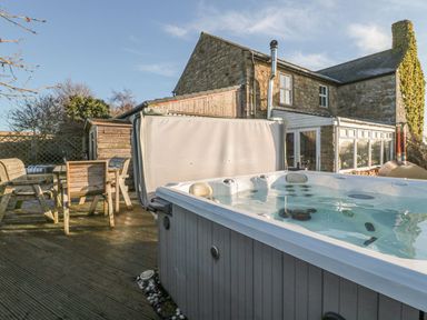 An outdoor patio with a hot tub and wooden table with chairs beside a stone house at Petty Knowes Cottage Rochester near Otterburn