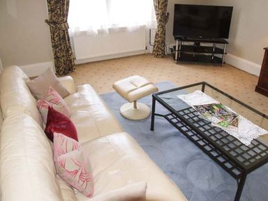 A living room with a cream sofa and patterned cushions a glass coffee table with magazines a cream ottoman a television on a stand floral curtains and beige carpet at Bank View in Chirnside