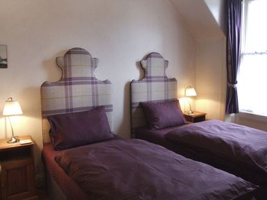 A bedroom with two single beds with plaid headboards and purple bedding two bedside tables with lamps and a window with purple curtains at Bank View in Chirnside