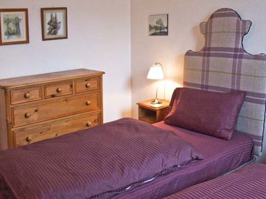 A bedroom with two single beds covered in purple bedding a wooden chest of drawers a nightstand with a lamp and framed artwork on the walls at Bank View in Chirnside