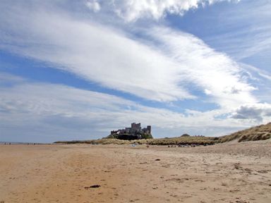 A sandy beach with grass-covered dunes and a castle in the distance under a partly cloudy sky