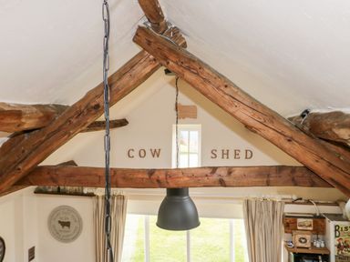 A room with exposed wooden beams and a hanging light above a window with curtains at Cowshed Normanby near Flyingthorpe