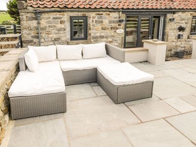 An outdoor patio with a gray wicker sectional sofa with white cushions next to a stone building at Cowshed Normanby near Flyingthorpe