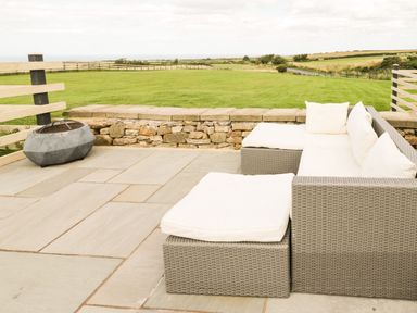 A patio with a cushioned wicker sofa and ottoman and a fire pit by a stone wall overlooking a grassy field at Cowshed Normanby near Flyingthorpe