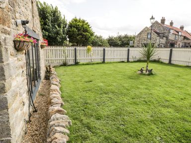 A backyard with a lawn a small palm tree stone wall and hanging flower baskets at Cowshed Normanby near Flyingthorpe