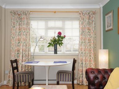 A dining area with a white oval table and two striped chairs by a window with floral curtains and a vase of flowers on the windowsill at Alway in Lifton