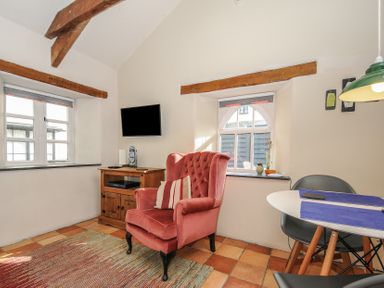 A sitting area with a pink armchair a wooden cabinet and a wall-mounted tv near windows and a table with chairs at The Barn in Lifton
