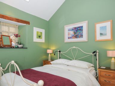 A bedroom with a double bed white bedding and a maroon runner two wooden bedside tables with lamps green walls and framed pictures at The Barn in Lifton