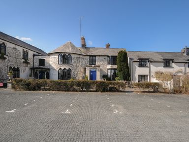 A photograph of a building with white walls and black framed windows behind a parking area at The Barn in Lifton
