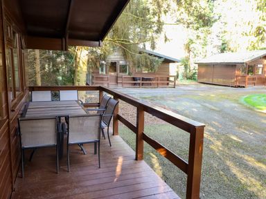 An outdoor wooden deck with a table and six chairs next to two wooden cabins surrounded by trees at Partridge in Withern near Alford