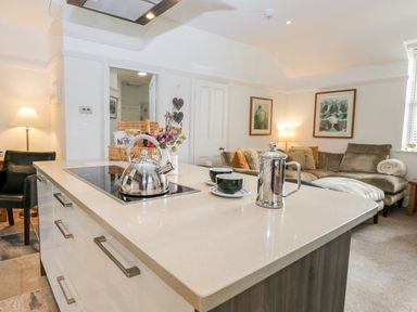 A kitchen island with a kettle, coffee pot, and cups with a living area with a sofa and framed pictures at The Bolt Hole in Kendal