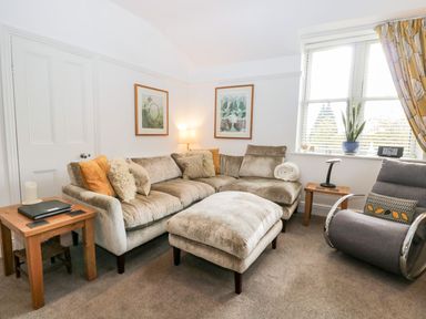 A living room with a beige sectional sofa and an ottoman a gray armchair wooden side tables and framed pictures on the wall at The Bolt Hole in Kendal
