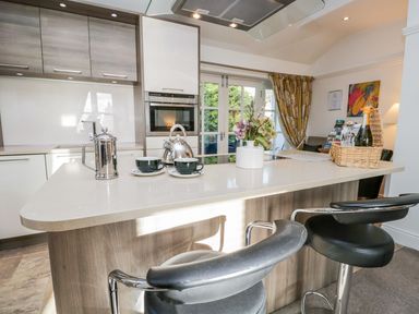 A kitchen with an island counter with coffee cups a kettle a flower vase and two bar stools at The Bolt Hole in Kendal