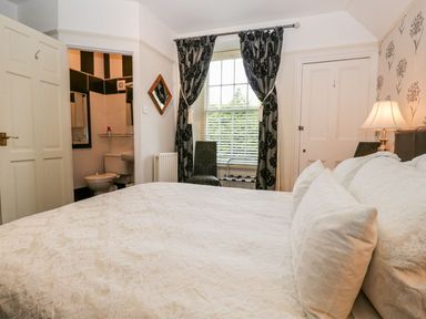 A bedroom with a bed white bedding a window with curtains a chair a bedside lamp and an en suite bathroom at The Bolt Hole in Kendal