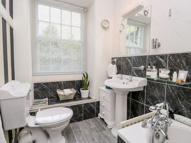 A bathroom with a toilet, pedestal sink, bathtub, window with blinds, small cabinet, and black and white tile walls at The Bolt Hole in Kendal