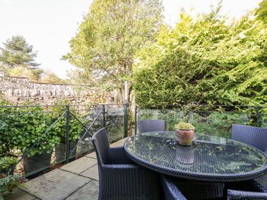 An outdoor patio area with a glass round table and four wicker chairs surrounded by greenery and a stone wall at The Bolt Hole in Kendal