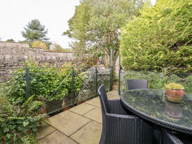 An outdoor patio area with a glass table, woven chairs, potted plants, stone wall, and greenery at The Bolt Hole in Kendal